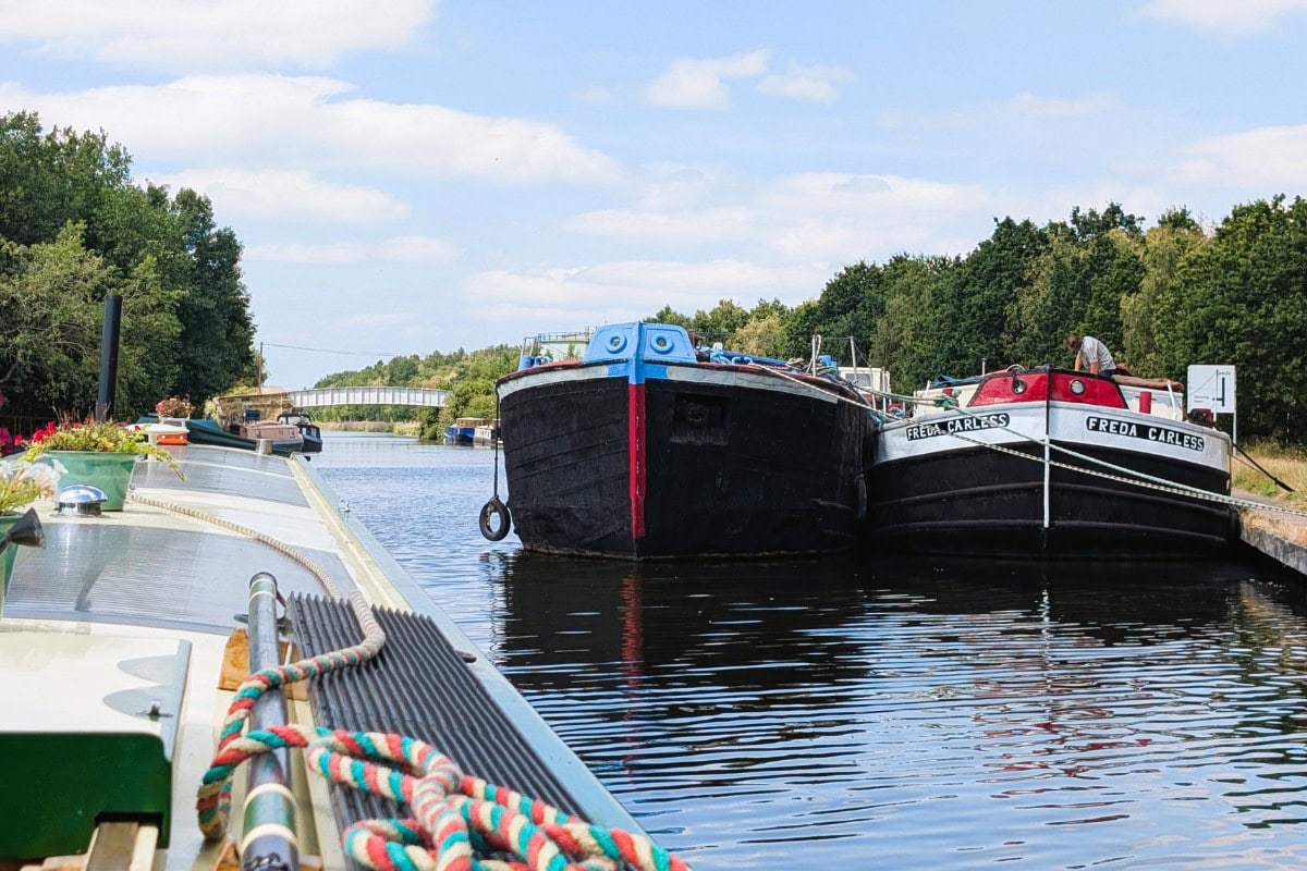 Narrowboats moored on a peaceful canal with lush green trees and blue sky.