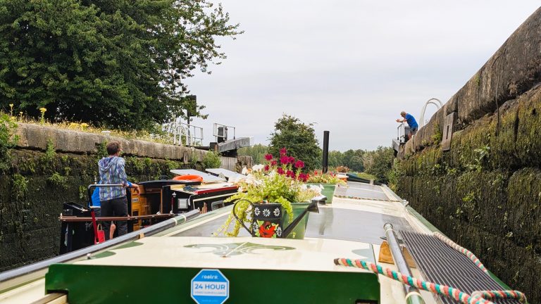 Narrowboat navigating a canal lock with people onboard and lush greenery.