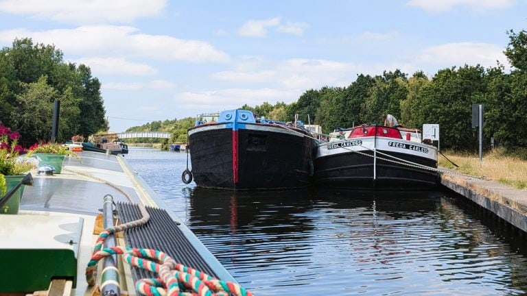 Two narrowboats moored on a peaceful canal with lush greenery and blue sky.
