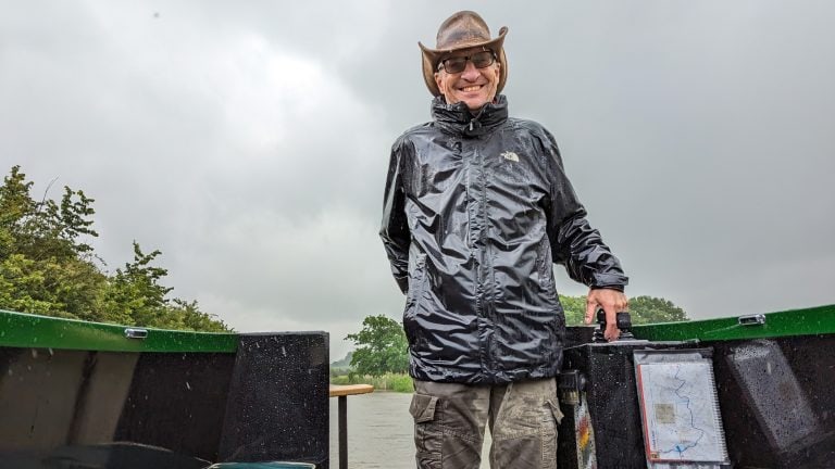 Smiling man in waterproof jacket on narrowboat during rainy day.