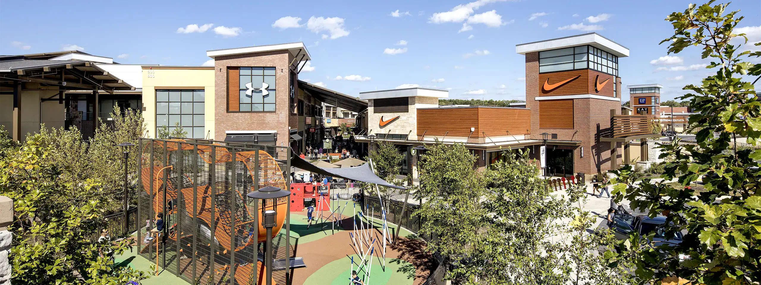 Outdoor shopping area with stores, trees, and a playground under a blue sky.