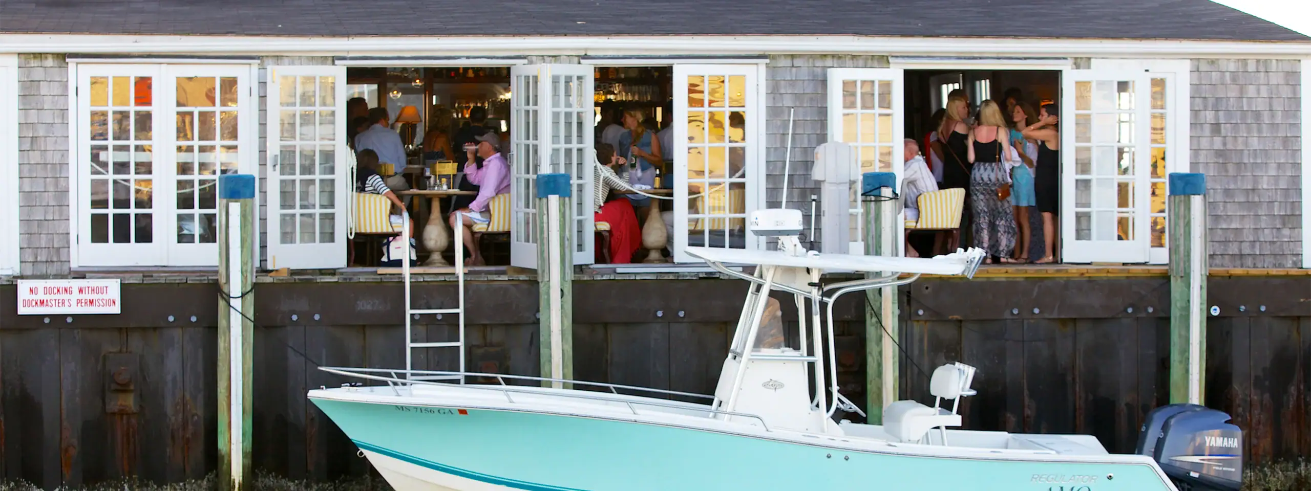 People dining inside a waterfront restaurant with large windows and a boat docked outside.