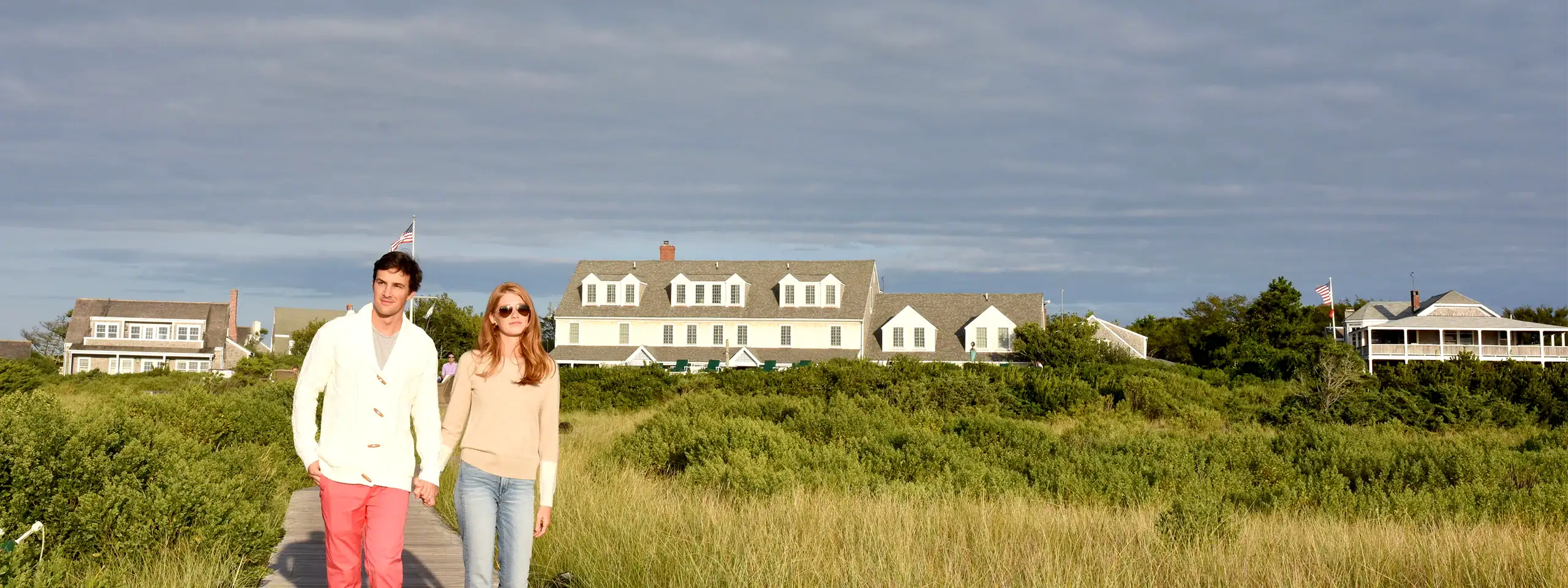 A couple walking on a grassy area near beach houses under cloudy sky.