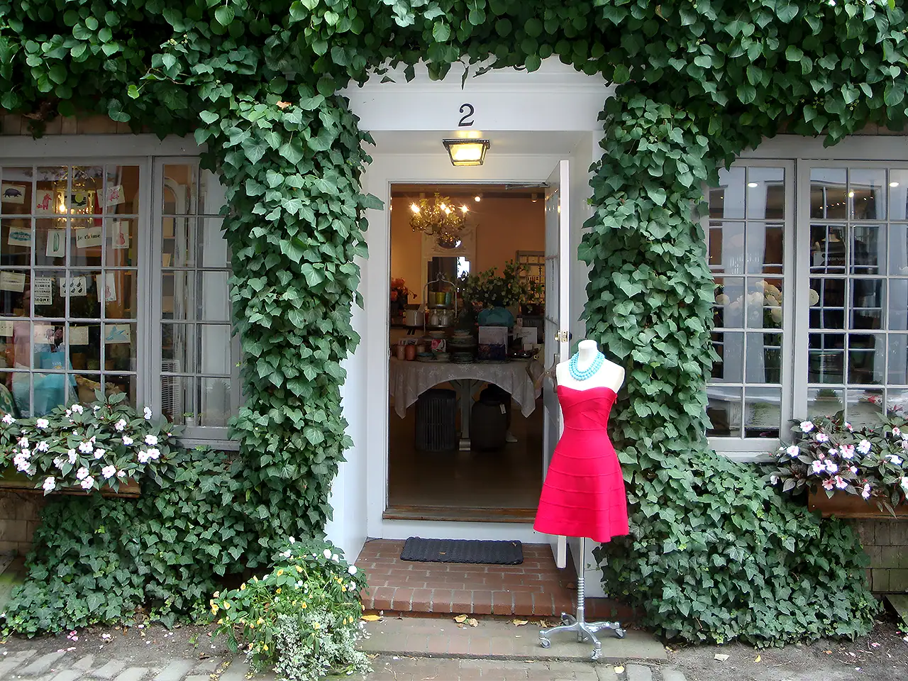Store entrance framed by lush green ivy and flower boxes, with a red dress on a mannequin outside.