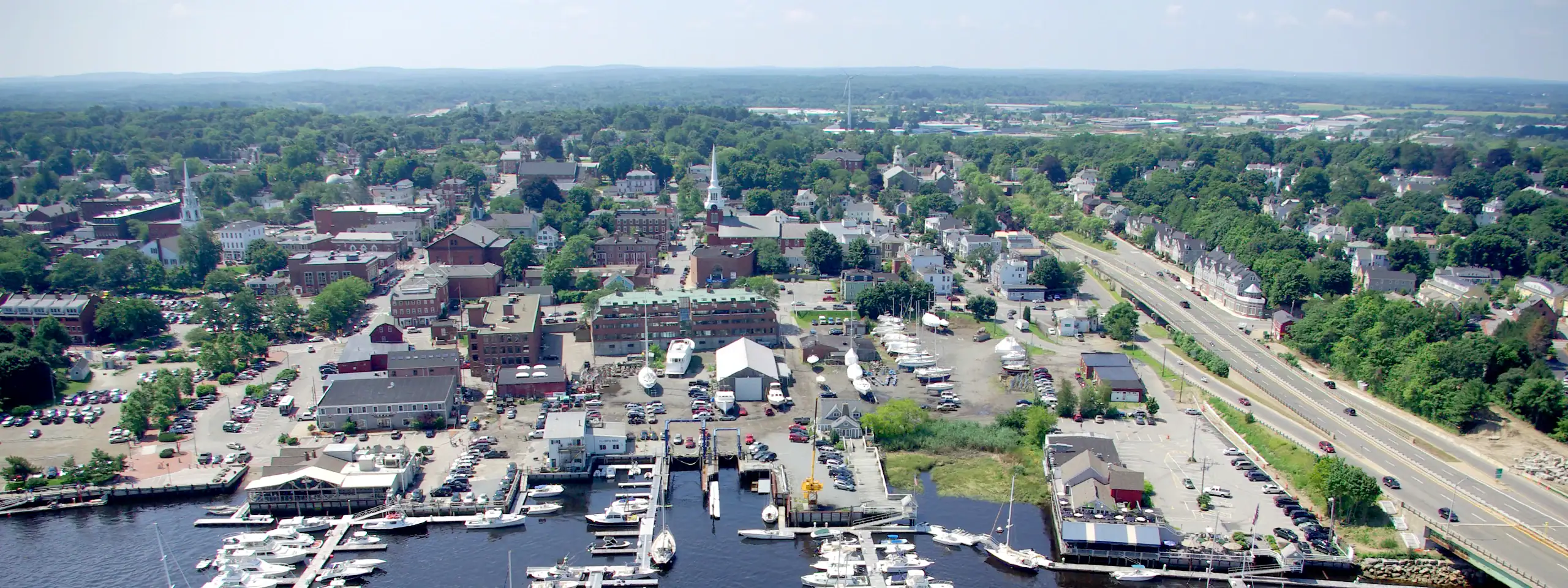 Aerial view of a city with a marina, parking lots, and green areas, near a highway.