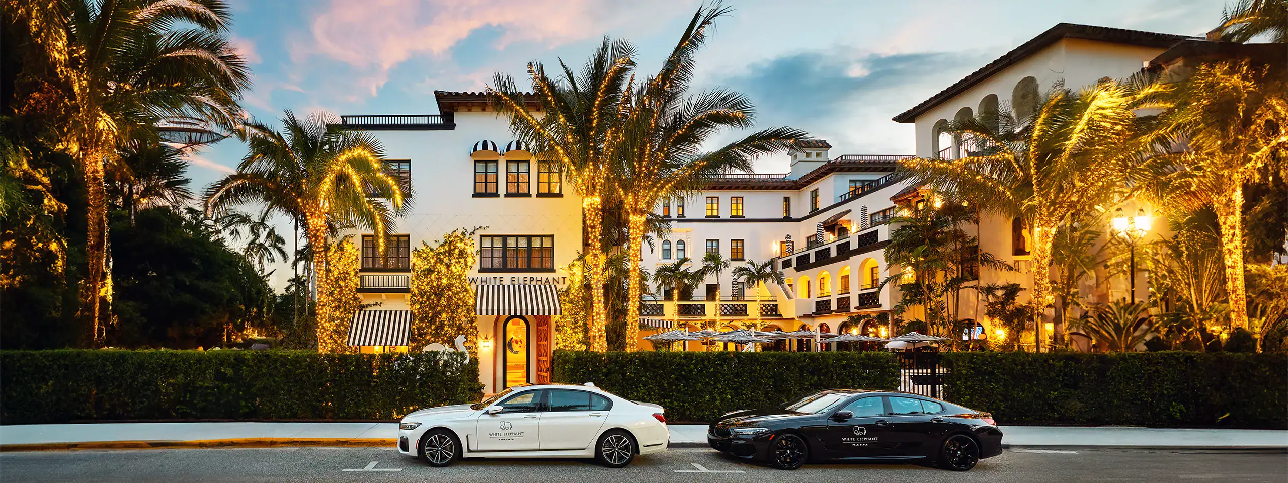 Luxury hotel with palm trees and cars in front at sunset.