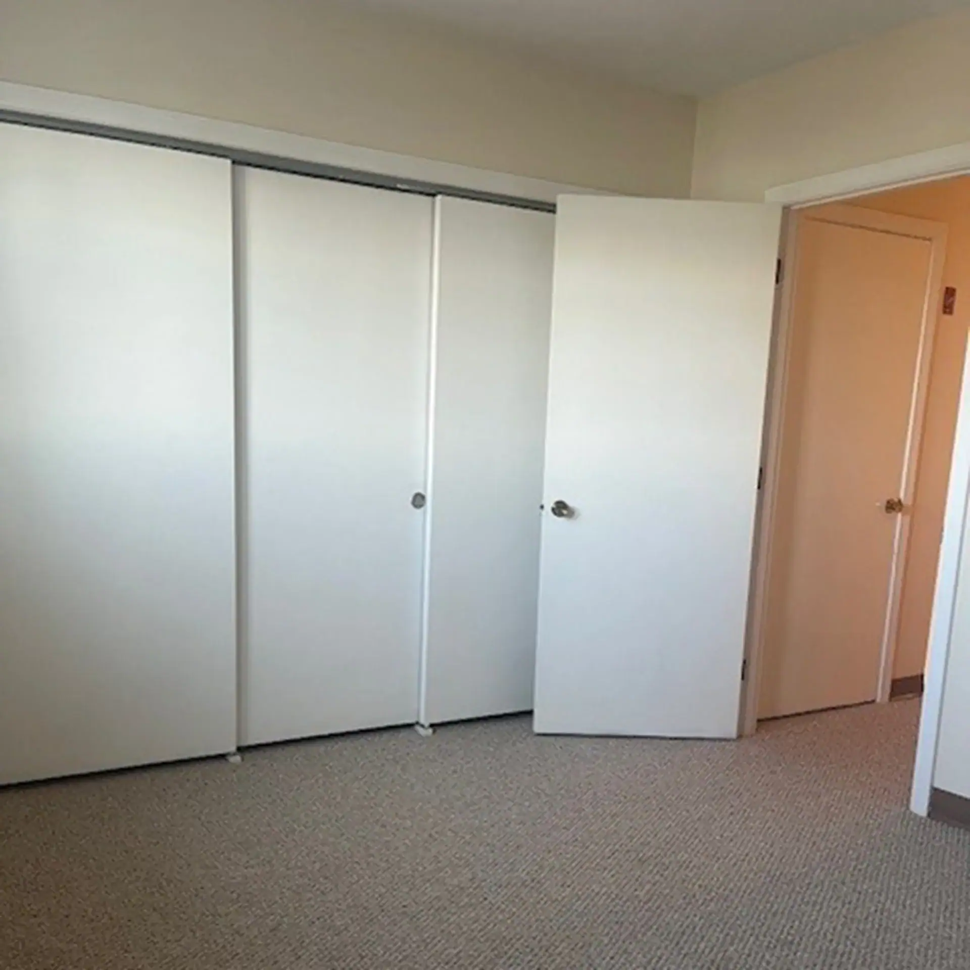 Empty room with white closet doors and an open door leading to another room.