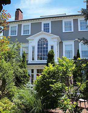 Large gray house with white trim and a prominent arched window, surrounded by lush greenery.