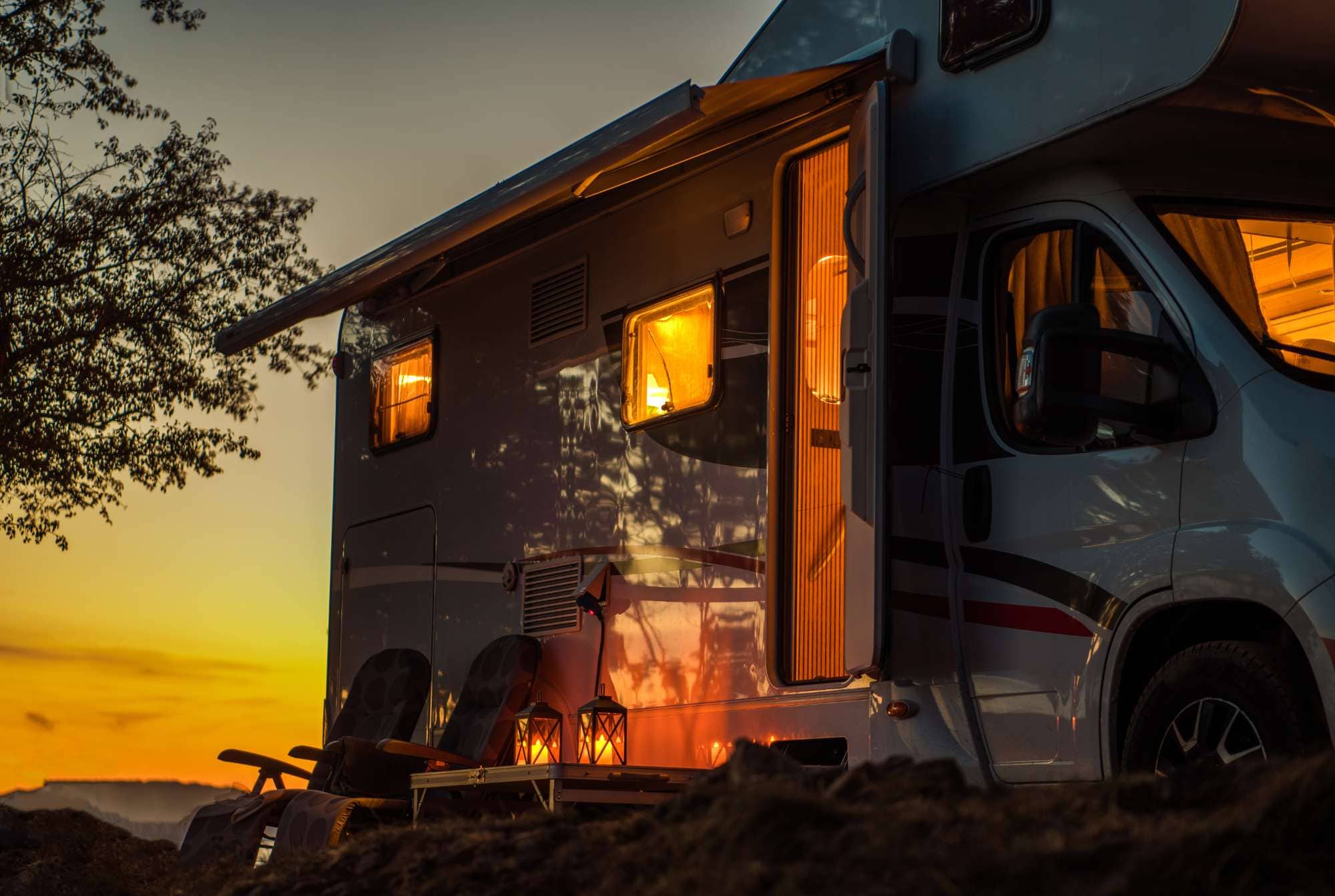Luxury RV parked at sunset with warm interior lighting and outdoor seating.