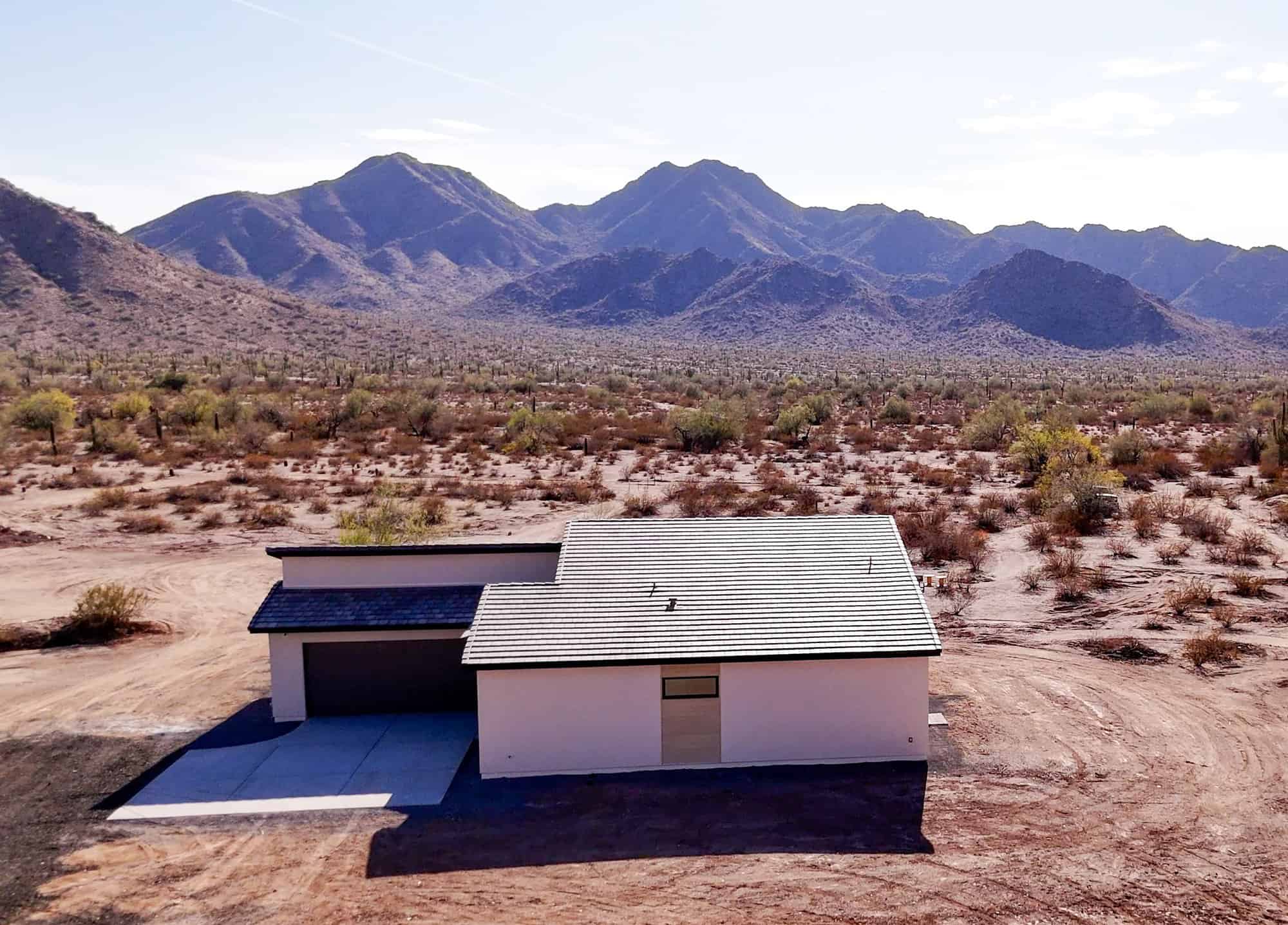 An aerial view shows a modern, single-story home with a white exterior and a dark, sloped tiled roof, situated in a vast desert landscape. The house features a dark garage door and a minimalist design with a single small, rectangular window visible on the rear wall. The surrounding terrain is arid, filled with sandy soil, low-lying desert shrubs, and cacti, all leading toward a backdrop of rugged, sun-drenched mountains under a clear sky.