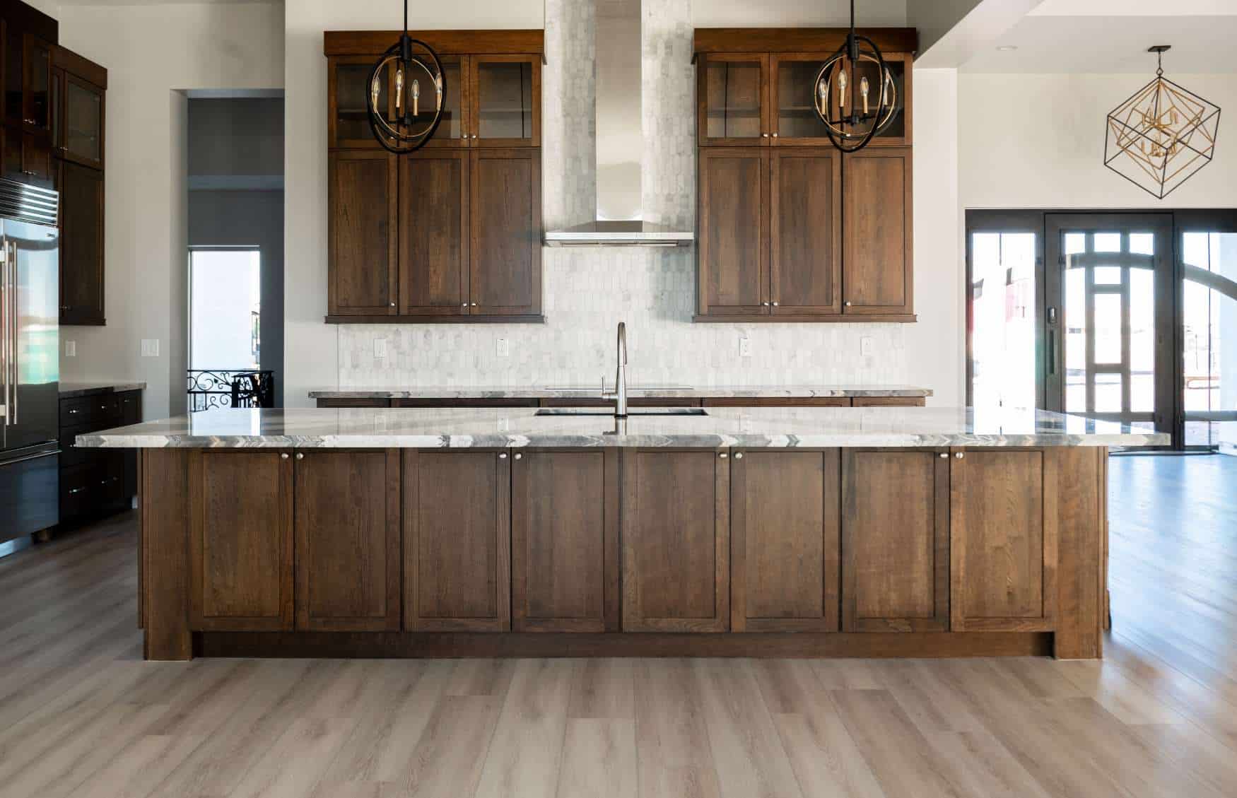 A wide shot captures a spacious, high-end kitchen centered around a massive island with dark wood cabinetry and a polished marble countertop. A stainless steel sink with a modern faucet is installed in the middle of the island. In the background, two sets of dark wood upper cabinets with glass-fronted tops frame a sleek stainless steel range hood, which is set against a white tiled backsplash. Two unique, circular black chandeliers hang over the island, while a geometric gold light fixture is visible in the adjacent room to the right. To the far left, a stainless steel refrigerator is partially visible, and the entire space features light-toned wood flooring.