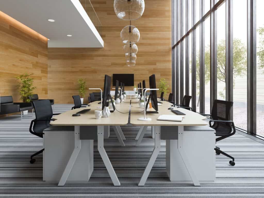 Two long, light-wood communal desks run parallel down the center of the room, equipped with black computer monitors, white task lamps, and ergonomic black mesh chairs.