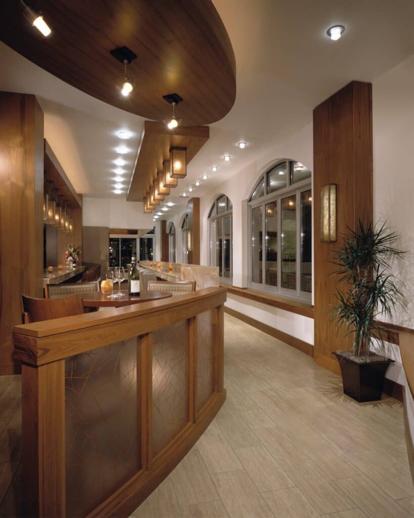 A low wooden partition with textured glass panels separates a hallway from a dining area. Behind the partition, a round table is set with wine glasses and a bottle. A long bar counter with stools extends into the background.