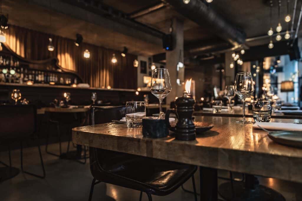 A low-angle, shallow-depth-of-field photograph captures a dimly lit, upscale restaurant interior. In the sharp foreground, a wooden dining table is set with clear glassware, a water tumbler, a small black container, and a dark pepper grinder adjacent to a flickering candle flame. Dark leather chairs are tucked around the tables. The background blurs softly into a warm haze of atmospheric lighting, featuring exposed Edison bulbs hanging over an elegant bar area on the left and various dining tables arranged throughout the industrial-chic space, which is characterized by exposed ceiling pipes and concrete pillars.