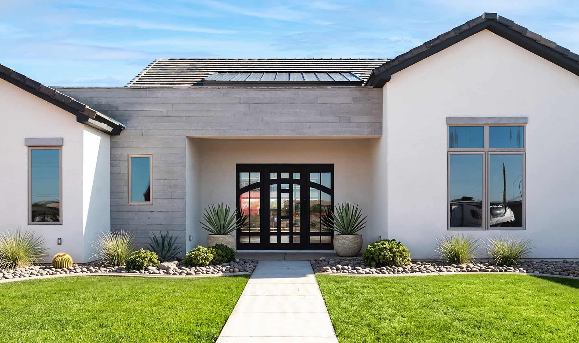 A medium shot focusing on the front entrance of a modern, desert-style home. The entryway is recessed beneath a flat, light-gray wood-planked overhang, featuring a large black-framed glass door with an arched window design. Flanking the entrance are two large, spherical stone planters containing agave plants. To the right, a prominent white stucco gabled section features a large three-pane window. The foreground shows a straight concrete walkway cutting through a vibrant green lawn, leading to the door. A landscaping bed of smooth river rocks, desert grasses, and small shrubs runs along the base of the house.