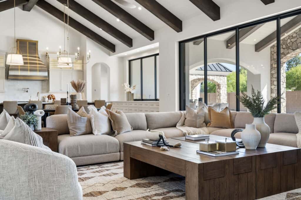 Nu Wavz Homes custom luxury open-concept living room featuring vaulted ceilings with dark wood exposed beams, a large neutral sectional sofa, a solid wood coffee table, and expansive sliding glass doors leading to an outdoor stone patio.