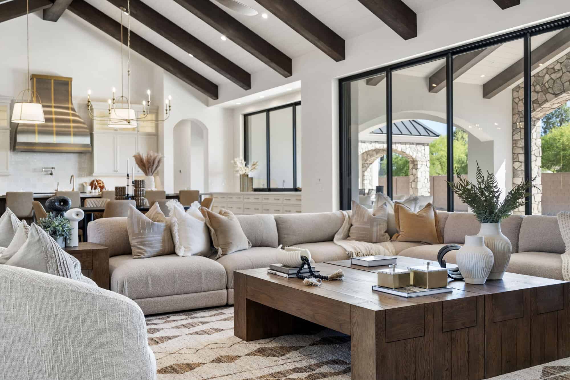 Nu Wavz Homes custom luxury open-concept living room featuring vaulted ceilings with dark wood exposed beams, a large neutral sectional sofa, a solid wood coffee table, and expansive sliding glass doors leading to an outdoor stone patio.