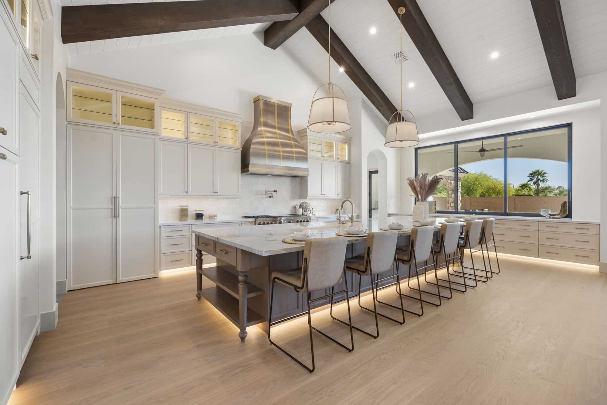 Luxury kitchen by NuWavz Custom Homes featuring vaulted ceilings with exposed wood beams, white custom cabinetry, a large quartz island with seating, stainless steel range hood, oversized picture window, and wood flooring.