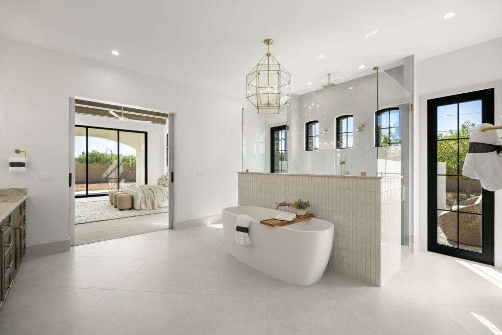 Nu Wavz Homes custom luxury primary bathroom design featuring a freestanding soaking tub with a wooden tray, a large geometric gold chandelier, a spacious glass-enclosed walk-in shower, and open sliding doors showing a view into the master bedroom suite.