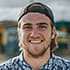 Smiling young man outdoors wearing a cap and patterned shirt.