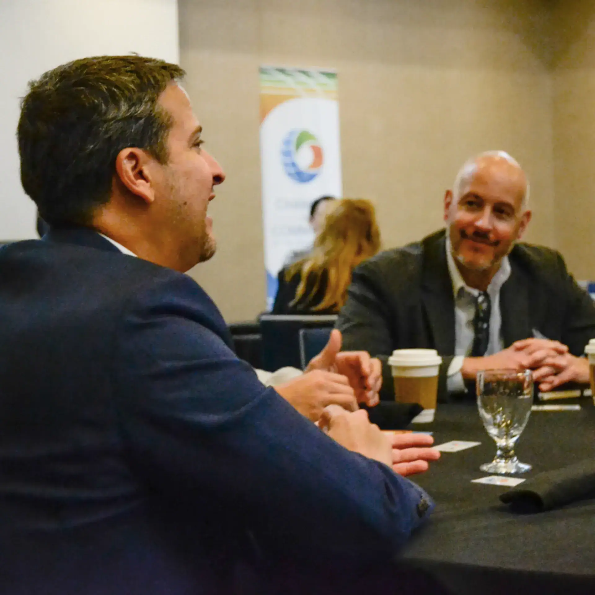 Two men in business attire talking at a professional event, with coffee and water on the table.