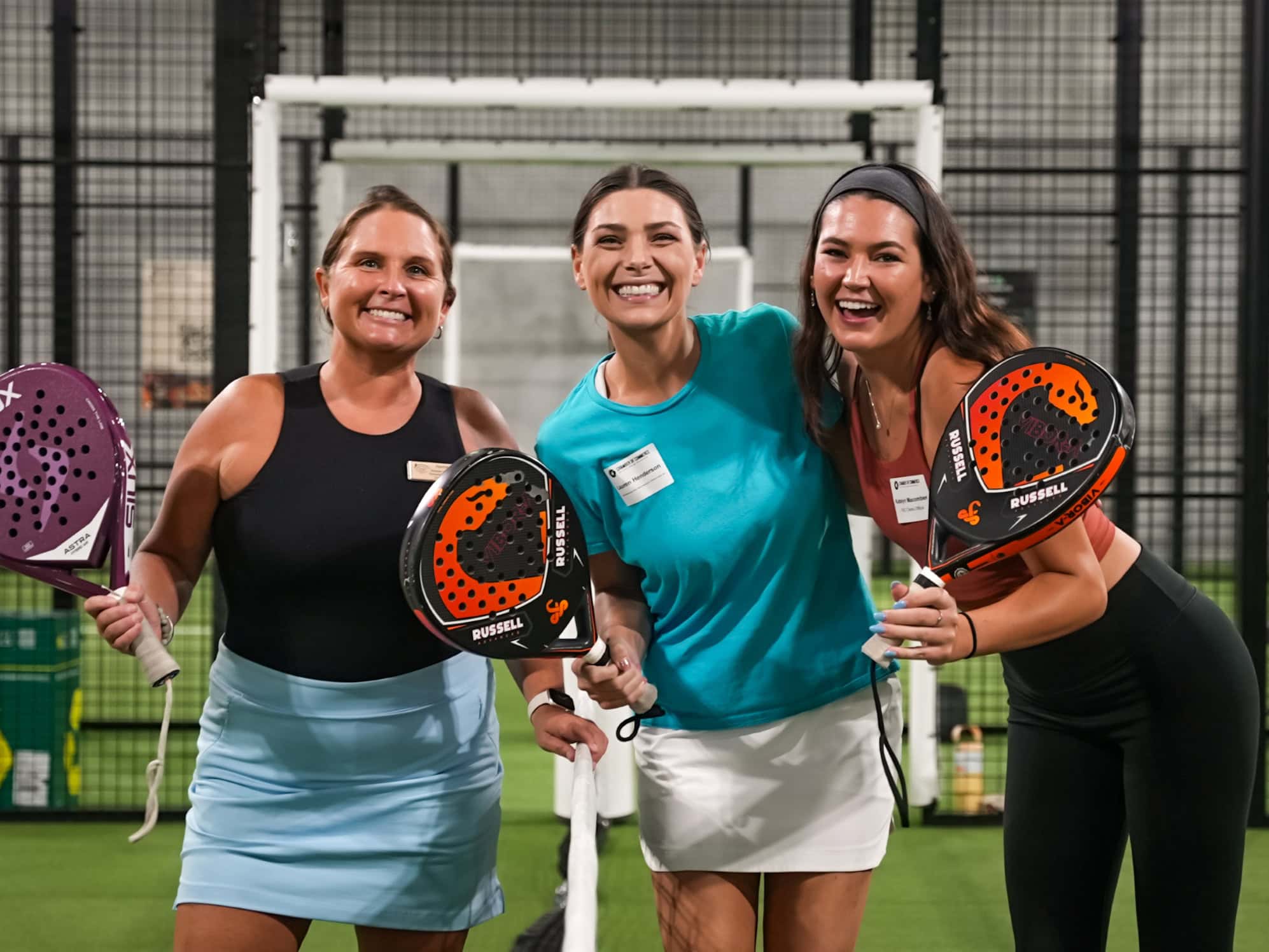 Three women smiling with paddle tennis rackets on indoor court.