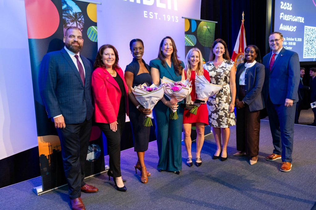 Group of seven diverse people on stage at awards event holding flowers.