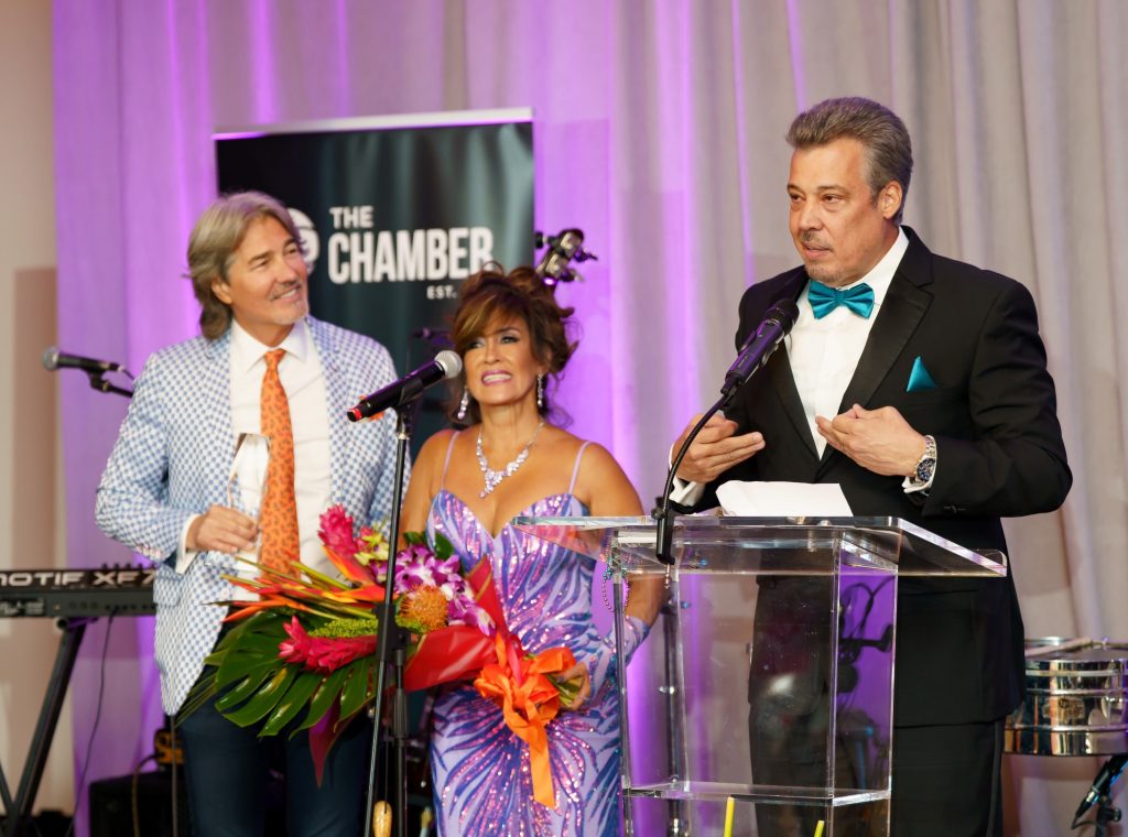Man in tuxedo speaking at a formal event with two women on stage.