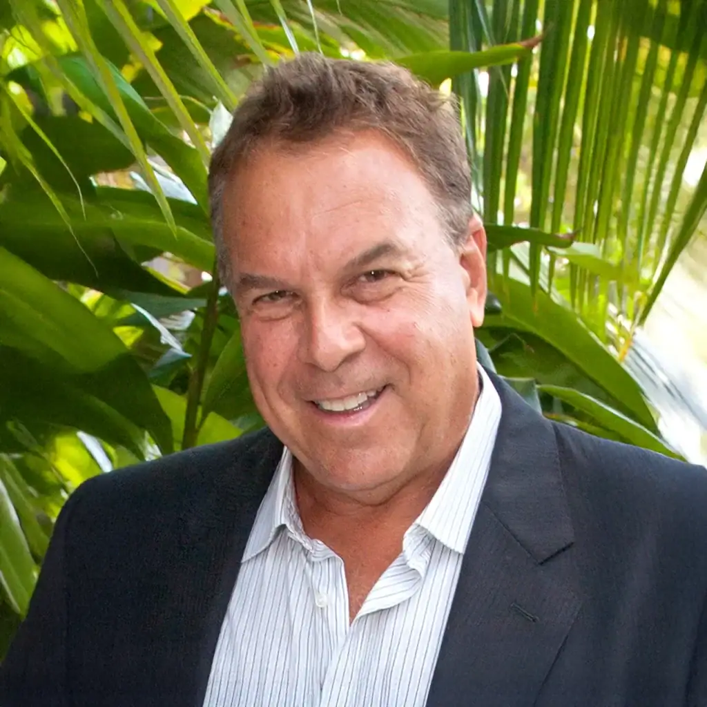 Smiling man in a suit outdoors with lush green foliage in the background.