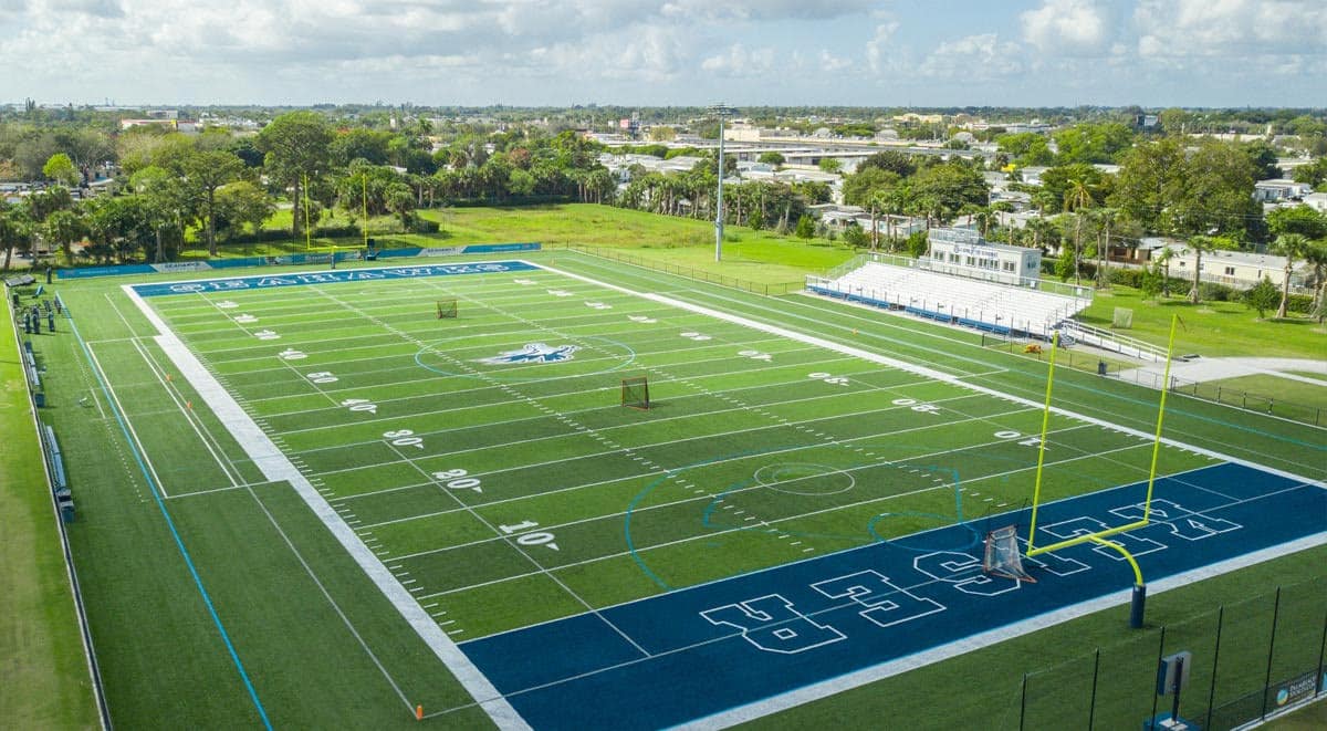 Aerial view of a large, well-maintained football field with blue end zones and surrounding greenery.