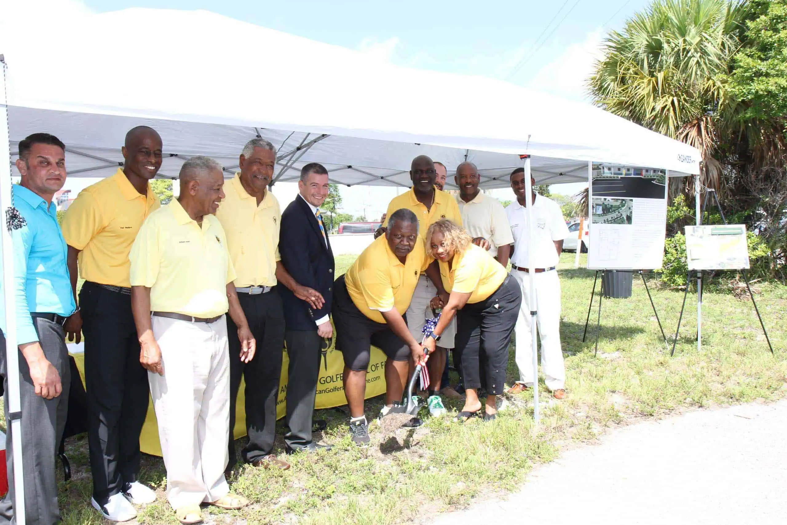 Group of people at golf event, some in yellow shirts, outdoors under a tent.