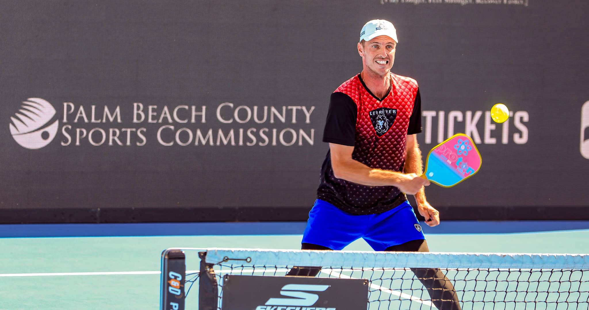 Player in red and black pickleball outfit prepares to hit ball at Palm Beach County Sports event.