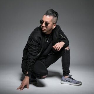 A young man in black casual attire crouching with sunglasses on, posing confidently for the camera.