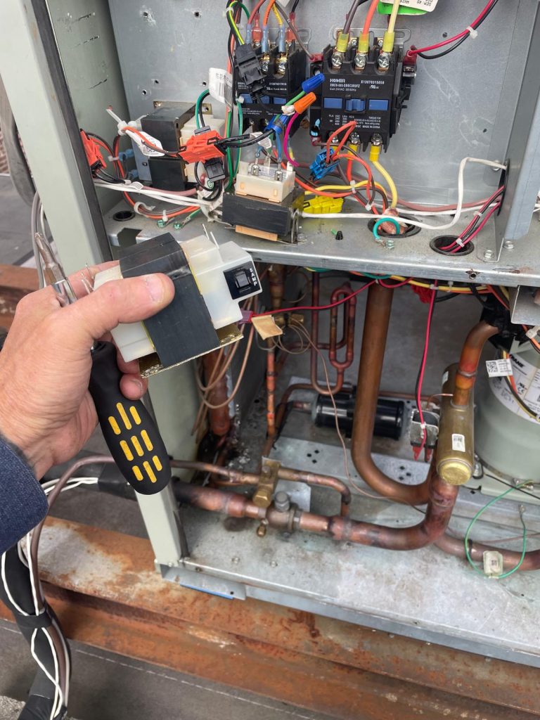 Hand holding an electrical component near an open HVAC unit with wiring.