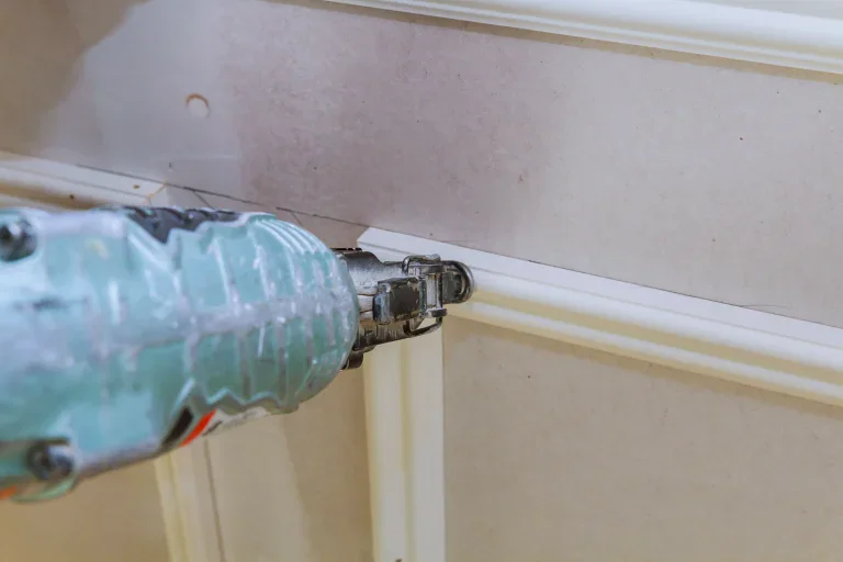 Close-up of a nail gun securing white crown molding to a wall.