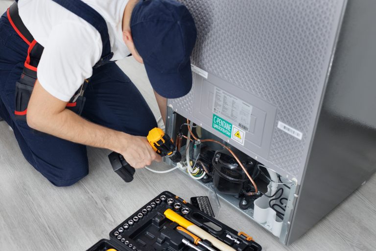 Man repairing the back of a refrigerator with a power drill.