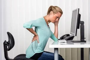 Back Pain Relief at Physio-Medicine London Woman experiencing back pain at her desk in an office setting.