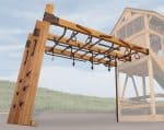 A wooden monkey bar structure with various rings and climbing holds is featured in a slightly low-angle shot. The structure is made of light-colored wood beams connected by dark metal brackets and includes multiple brown hand rings, triangular grips, and rope attachments. On the left, a slanted wooden wall with climbing pegs is visible. A thin metal pole supports the right side of the monkey bar section. In the blurred background, a grassy slope and a faint outline of a wooden playhouse can be seen, suggesting an outdoor play area.