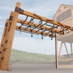 A wooden monkey bar structure with various rings and climbing holds is featured in a slightly low-angle shot. The structure is made of light-colored wood beams connected by dark metal brackets and includes multiple brown hand rings, triangular grips, and rope attachments. On the left, a slanted wooden wall with climbing pegs is visible. A thin metal pole supports the right side of the monkey bar section. In the blurred background, a grassy slope and a faint outline of a wooden playhouse can be seen, suggesting an outdoor play area.