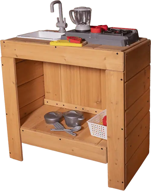 A wooden toy kitchen unit is shown from a slightly elevated angle. The unit has a light wood finish and an open-shelf design. On the top surface, from left to right, there's a white plastic sink with a gray faucet, a yellow cutting board with a red watermelon slice, and a gray stovetop unit with red burners. A gray blender sits behind the stovetop. The open shelf below holds two gray toy pots with lids, a gray toy spatula and ladle, and a white plastic basket containing a red item.