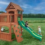 Wooden outdoor playset with slide and climbing wall for kids in a grassy yard.