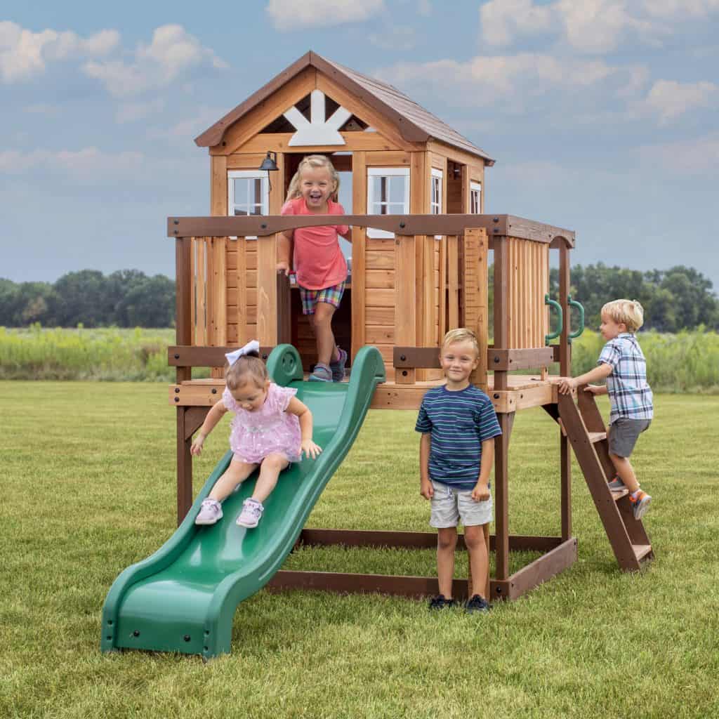 Children playing on a wooden playhouse with slide in a grassy field.