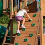 Young girl climbing on a wooden outdoor playset with green handholds.