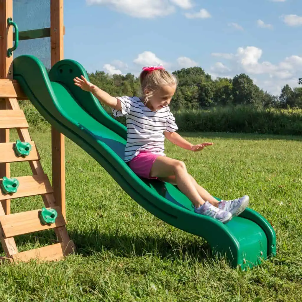 A young girl with a pink bow in her hair smiles with her arms outstretched as she slides down a wavy green plastic slide. She is wearing a black and white striped shirt, pink shorts, and purple sneakers.