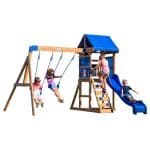 A wooden children's playset with bright blue accents is isolated against a solid white background, featuring four children at play. On the left, a boy and a girl are using two blue belt swings attached to a sturdy wooden beam. In the center, a girl in a pink shirt climbs a wooden ladder equipped with blue rock-climbing holds to reach the upper clubhouse, which is covered by a blue fabric roof and enclosed with blue mesh safety walls. To the right, a boy in a dark t-shirt slides down a wavy blue plastic slide. The structure is built with light-colored wood and blue hardware.