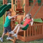 A medium shot of two children playing on a wooden backyard playset. A young girl with blonde hair in a ponytail and a red t-shirt is seated on a green belt swing, holding onto green plastic-coated chains. To her left, a young boy in a green and gray raglan shirt and dark shorts climbs a wooden ladder toward the playhouse platform, reaching one hand out toward the girl. The playset is constructed from light brown cedar and features a green slide, green safety handles, and a lower-level sandbox or play area enclosed by wooden slats. The scene is set on a lush green lawn during a bright, clear day.