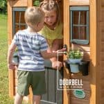 A young boy in a striped shirt reaches out to press a red doorbell button on the front of a wooden playhouse. A young girl in a yellow shirt watches him from just inside the open doorway. The playhouse features light-colored wood paneling, dark-framed windows, and a white potted plant sitting in a black window box. White text overlaid on the image reads "DOORBELL WITH CHIME," with a line pointing directly to the doorbell hardware. The scene is set outdoors on a sunny day with green grass visible in the background.