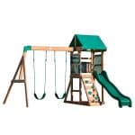 A wooden children's playset with forest green accents is shown against a solid white background. The structure features a main tower with a green fabric gabled roof, an upper deck enclosed by green mesh safety walls, and a built-in chalkboard. Attached to the tower is a wavy green slide on the right and a wooden ladder with green rock-wall grips in the center. To the left, a swing beam holds two green belt swings with green-coated chains. The entire playset is constructed from light-toned wood with green metal brackets and hardware.