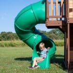 Children playing on a green outdoor slide attached to a wooden playset.