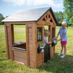 Two young children are playing with a small wooden playhouse situated on a sunny, grassy lawn. The playhouse features a dark shingled roof, a half-door, and window boxes. A young girl with a blue bow in her hair stands outside, tending to a small potted plant in one of the window boxes. A young boy is visible peeking out from behind the door, while a toy kitchen set with a sink and stovetop is visible through the large side opening of the structure. The background shows a wide, open field under a clear blue sky.